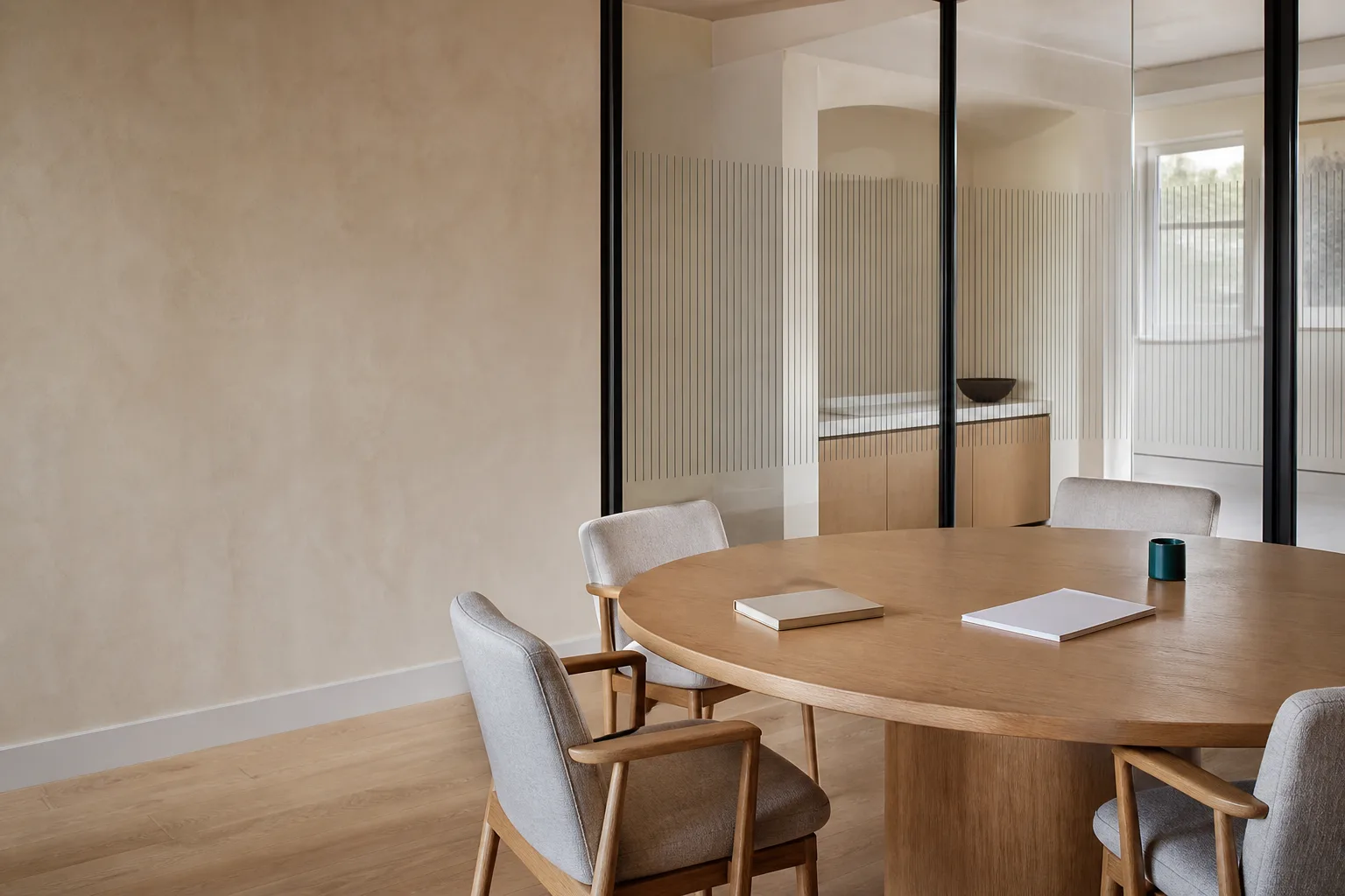 A quiet meeting room with a round oak table, four upholstered chairs, two closed notebooks, a teal cup, and a slatted glass partition in soft daylight.